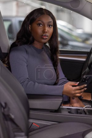 The image captures a young woman seated in the drivers seat of a car, engaging with a smartphone in her hands. Her focused gaze and the careful handling of the device suggest she is attentive to the