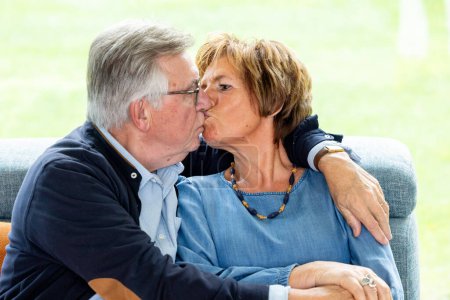 This image features an elderly couple in a loving embrace, sharing a gentle kiss. The man, with silver-grey hair and glasses, kisses his partner, who wears a blue blouse and a beaded necklace. They