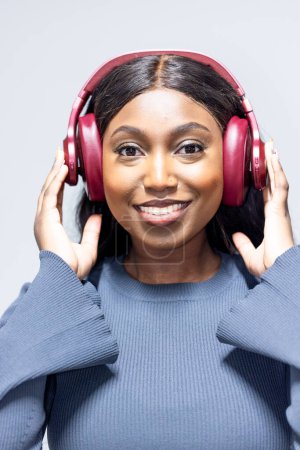 This is a portrait of a cheerful young African woman with black hair, wearing a blue long-sleeved top and adjusting her red headphones. The headphones are clearly a significant feature, indicating a