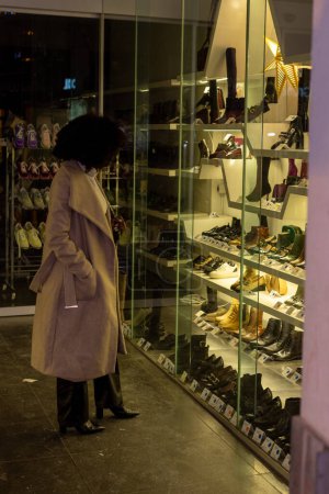 This image captures a woman engaged in the timeless activity of window shopping. Standing outside a shoe store, she appears absorbed by the display, her silhouette reflected in the glass. The warm