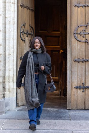 Una joven mujer emerge de la gran puerta de una catedral, mezclando la moda contemporánea con la elegancia atemporal de la arquitectura gótica. Su salida equilibrada sugiere una narrativa de exploración o