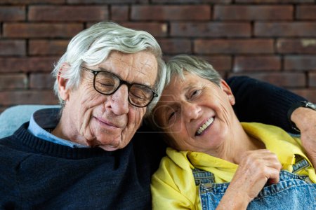 Téléchargez les photos : Une image tendre montrant un couple de personnes âgées dans un moment de joie et de contentement. Le monsieur, avec ses cheveux argentés et ses lunettes, a les yeux doucement fermés, souriant sereinement. La dame, arborant un jaune - en image libre de droit