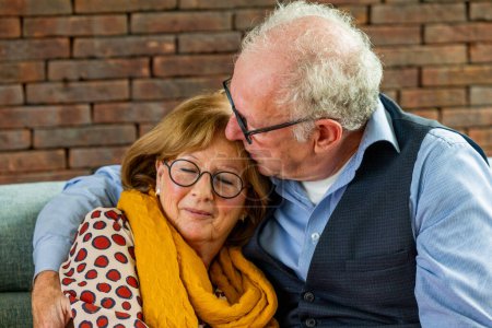In this intimate scene, an elderly man with grey hair and glasses is depicted giving a tender kiss on the forehead to his partner. The woman, wearing stylish glasses and a scarf, closes her eyes and