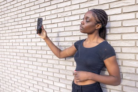The image depicts a young African woman engaged in taking a selfie, holding her smartphone up with one hand. She stands in profile to the camera, with a white brick wall providing a uniform and