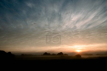 As the day begins, a mystical dawn breaks over fields veiled in mist, with the sun peeking through the low-lying fog, hinting at the landscapes hidden contours. The sky above is a canvas of dramatic