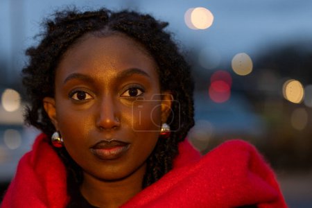 This portrait captivates with a young womans direct stare into the lens, her face illuminated by the gentle dusk light. The red scarf is a vibrant contrast to the twilight, draped around her for