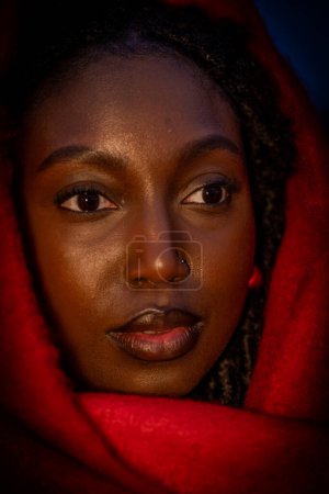 In this intimate close-up, the subjects face is partially shrouded in shadow, illuminated intriguingly by ambient light that highlights the contours of her face. The crimson fabric of her wrap