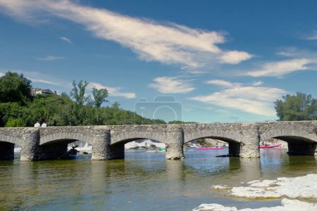 This vibrant and sunny scene captures the essence of summer in the Ardeche region, with individuals leisurely canoeing under a majestic old stone bridge that crosses the calm river. The bridge, with