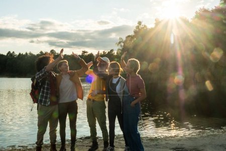 A vibrant multiracial group of young friends is captured in a moment of celebration by the lake, with the setting sun casting a glorious backdrop. Their postures suggest a cheer or a toast to the