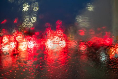 This vibrant image is taken from the perspective of a driver inside a vehicle during a rainy night. The scene is illuminated by the red bokeh of traffic lights, which are beautifully diffused by the