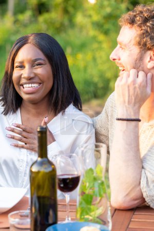 Esta imagen muestra un momento encantador entre dos amigos disfrutando de una noche de vino al aire libre. La mujer se ríe de todo corazón, con la mano en el pecho, mientras el hombre sonríe, mirándola con una