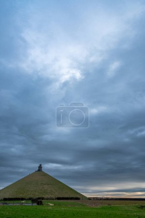Photo for This image features the iconic Lions Mound at Waterloo, with its monumental earthwork and the regal lion statue atop, against a dramatic overcast sky. The mound rises distinctly from the flat - Royalty Free Image