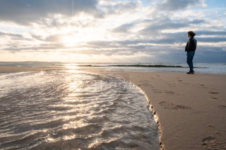 Cette image capture avec éloquence un individu solitaire debout sur la plage, regardant vers la mer sous la douce lumière du soleil couchant. La réflexion des soleils sur l'eau crée un chemin de lumière