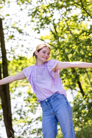 The image portrays a youthful woman mid-dance, her arms outstretched as if to welcome the embrace of a summer breeze. She wears a lilac-colored top, complementing the lively greens of the trees in the