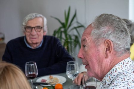 A senior man with a surprised look on his face adds a lively dynamic to a dinner party, as he listens to a story or comment from across the table. Another man, in the background, wearing glasses and a