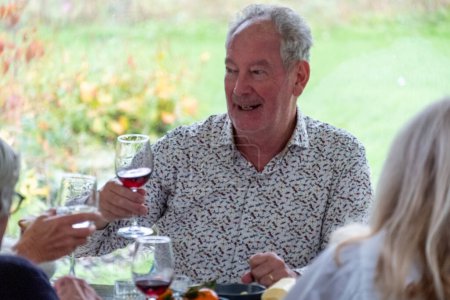 A joyful senior man with a glass of red wine is captured mid-toast, sharing a light-hearted moment with friends at a gathering. His expressive face and open smile convey the pleasure and warmth of the