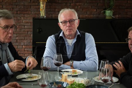 A senior gentleman with glasses and wearing a smart vest over a long-sleeve shirt gazes directly at the camera, offering a moment of contemplation amidst a lively dinner party. To his left, another