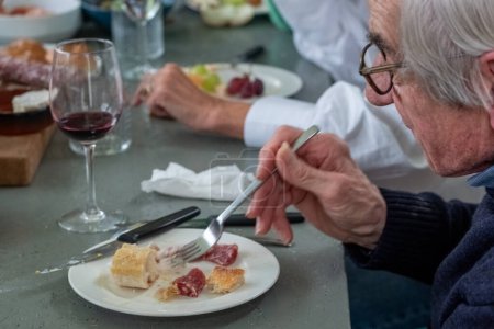 Un homme âgé avec des lunettes est vu profiter d'une belle sélection de fromage et de charcuterie, associée à un verre de vin rouge. Son accent sur les saveurs reflète un palais éclairé, et l'intime