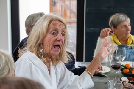 Mitten im Gespräch zeigt sich eine ältere Frau mit blonden Haaren und weißer Bluse überrascht oder aufgeregt. Ihre lebhafte Geste und ihr offener Ausdruck deuten auf eine lebhafte Diskussion hin oder