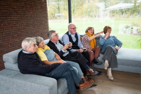 A delightful scene unfolds as a group of seniors gather on a sofa for a game day. Some hold game controllers, fully engaged in the playful competition, while others cheer and laugh, sharing in the fun