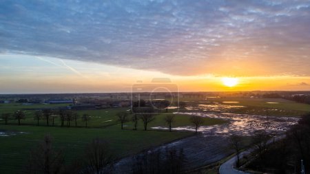 The image portrays a breathtaking sunset over a rural landscape with wetlands. The setting sun casts a warm glow across the water, highlighting its presence amidst the fields. A curving road in the