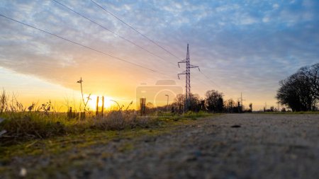 This image presents a ground-level view of a country road at sunset, with the focus on the power lines against the warm glow of the setting sun. The low angle emphasizes the rough texture of the road