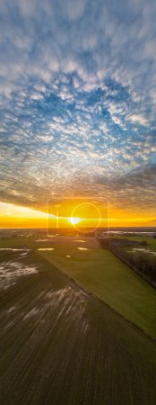 This vertical portrait captures the grandeur of a sunset descending over farmlands. The sky, filled with a blanket of textured clouds, is aflame with the golden hues of the setting sun. Below, the