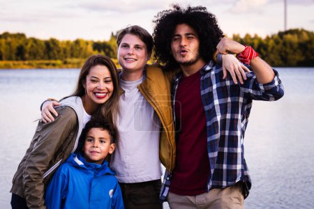 This vibrant image showcases a diverse group of friends and a young child enjoying a moment together by a lake. The group is composed of a woman with a bright smile, a young boy in a blue jacket
