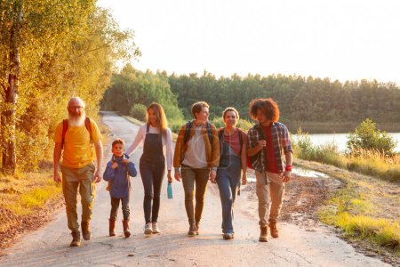 Una conmovedora escena de camaradería multigeneracional se representa en esta imagen, donde un grupo de amigos disfrutan de un tranquilo paseo por un sendero rural junto al lago mientras se pone el sol. El grupo es diverso