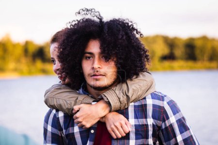 This image captures a young couple in an intimate embrace by the water, with a sense of introspection and closeness. The man in the foreground, with curly hair and a plaid shirt, gazes into the