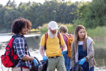 This image captures a diverse group of individuals participating in an environmental cleanup by a lake. A young man with curly hair and a plaid shirt holds a trash bag, alongside an older man with a