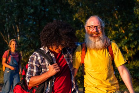 This image depicts a lively moment among friends in an outdoor setting at dusk. The focus is on two men in the foreground: a younger man with curly hair leaning in and an older man with a white beard