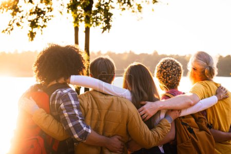 La imagen captura a un grupo de amigos en un abrazo cercano, mirando hacia fuera sobre un lago sereno como el día llega a su fin. El sol poniente arroja un cálido resplandor sobre la escena, simbolizando la comodidad y
