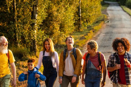 This image captures a group of friends from different generations enjoying a walk along a sunlit path surrounded by trees with autumn foliage. A young boy at the front looks confidently at the camera