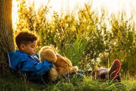 The image beautifully captures a young boy in a contemplative moment as he sits with his teddy bear against a tree, looking out over a lake bathed in the warm light of the setting sun. The boys