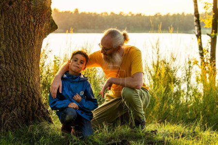 This image captures a tender moment between a grandfather and his grandson sitting by a tranquil lakeside at sunset. The elderly man, with a white beard, glasses, and a warm smile, gently places his