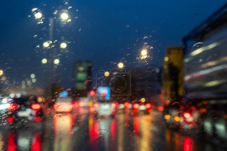 Dieses Bild zeigt den Blick eines Autofahrers durch eine regengesprenkelte Windschutzscheibe im abendlichen Berufsverkehr. Der Schein von Straßenlaternen und Fahrzeugheck erzeugt einen Bokeh-Effekt, mit dem kühlen Blau des