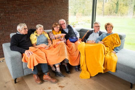 This image radiates comfort as a group of senior friends relax together on a couch, sharing a warm orange blanket. Their casual attire and the intimate setting create a scene of leisure and