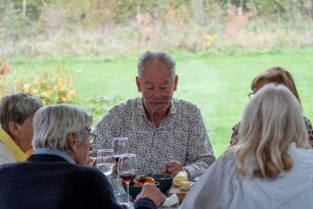Dieses Bild zeigt einen älteren Mann beim Essen mit Freunden. Er lächelt und scheint ein angenehmes Gespräch mit der ihm gegenüberliegenden Frau zu führen. Sie sitzen an einem Tisch, beladen mit