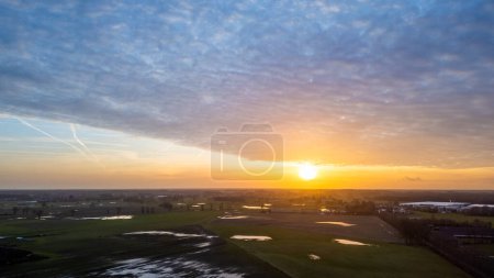 This aerial image captures the tranquil beauty of dawn as the sun breaks through a cloudy sky, casting a warm light over the wet fields below. The landscape is a patchwork of greens and dark