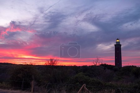 This striking image captures the silhouette of a towering lighthouse set against the fiery canvas of the sky at twilight. Vivid streaks of red and pink cut through the blue of the evening, as the