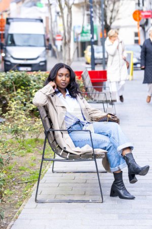 A young African woman with long dark hair sits casually on a metal bench in an urban park setting. She exudes a relaxed yet confident demeanor, dressed in a light blue shirt, cuffed blue jeans, and