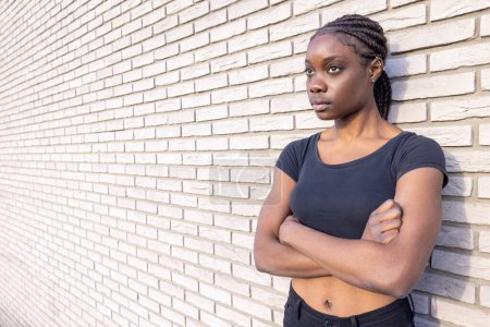Téléchargez les photos : Cette image représente une jeune afro-américaine aux cheveux tressés, bras croisés, appuyée contre un mur de briques blanches texturées. Elle porte un haut noir ajusté et regarde sur le côté avec une résolution - en image libre de droit