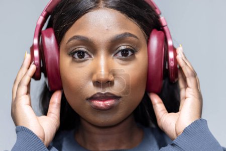 A close-up portrait of an African American woman, lost in her own world with deep red headphones that encompass her ears. Her hands are gently placed on the headphones, suggesting a careful adjustment