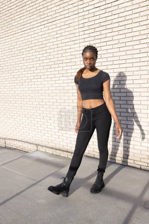La fotografía muestra a una joven africana de pie con confianza en un entorno urbano. El sol proyecta su sombra sobre una pared de ladrillo blanco texturizado, complementando su postura asertiva. Ella está vestida