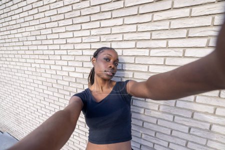 L'image capture une jeune femme africaine prenant un selfie avec un effet grand angle, étendant son bras vers la caméra. Sa pose est détendue mais expressive, avec une légère inclinaison de la tête et un calme
