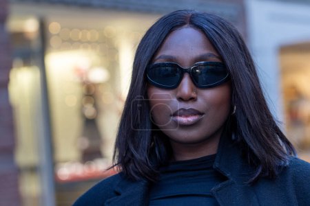 Esta imagen presenta a una mujer afroamericana adornada con un traje negro elegante, complementado con gafas de sol elegantes y oscuras que le dan un aire de enigma. La fotografía se toma al aire libre, con el bokeh suave