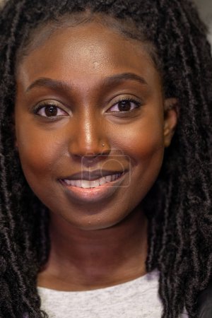 Das Foto zeigt eine junge schwarze Frau mit einem weichen und selbstbewussten Lächeln. Ihre natürliche Twist-Out-Frisur umrahmt ihr leuchtendes Gesicht perfekt und hebt ihre Augen hervor, die vor Lebendigkeit funkeln. Die