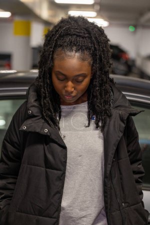 Esta imagen evocadora presenta a una joven mujer negra en contemplación, su mirada hacia abajo en un momento de introspección. Revestido con una práctica chaqueta negra y con capas sobre una camiseta gris relajada