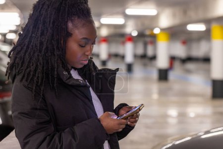 This image features a young woman absorbed in her smartphone within the confines of a sparsely populated parking garage. The soft gleam of the phone screen illuminates her concentrated face while her